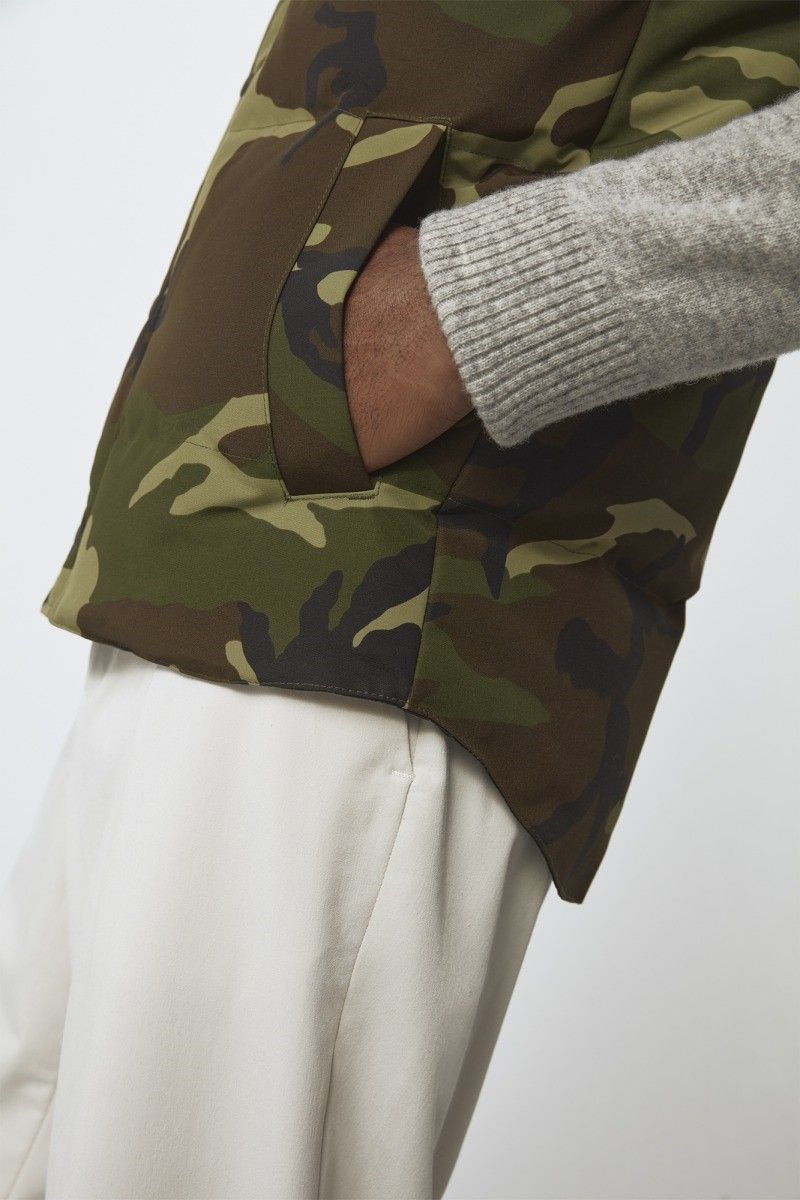 Close up of a person wearing a camouflage vest with a gray sweater and white pants, hand in the pocket.