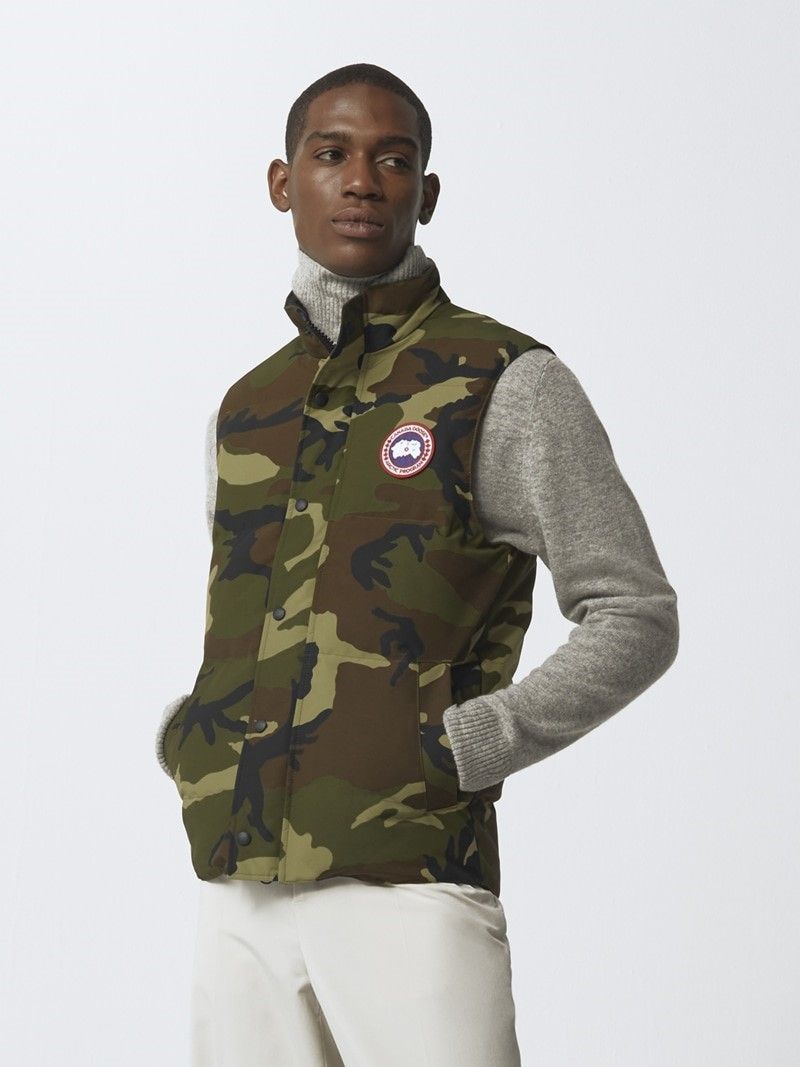 Man wearing camo vest with round logo on chest, gray turtleneck and white pants.
