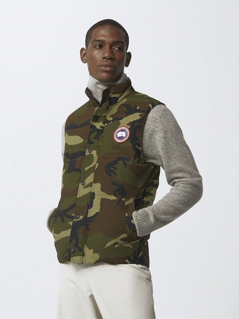 Man wearing camo vest with round logo on chest, gray turtleneck and white pants.