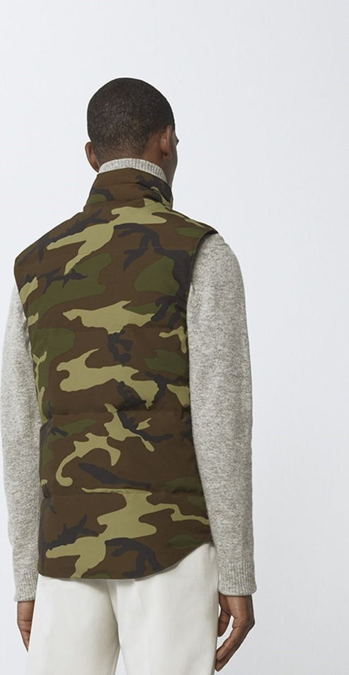 Man wearing a camo puffer vest over a gray turtleneck sweater with white pants.
