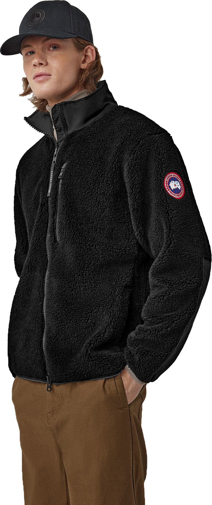 A person wearing a black fleece jacket with a circular logo on the left sleeve and a black cap.