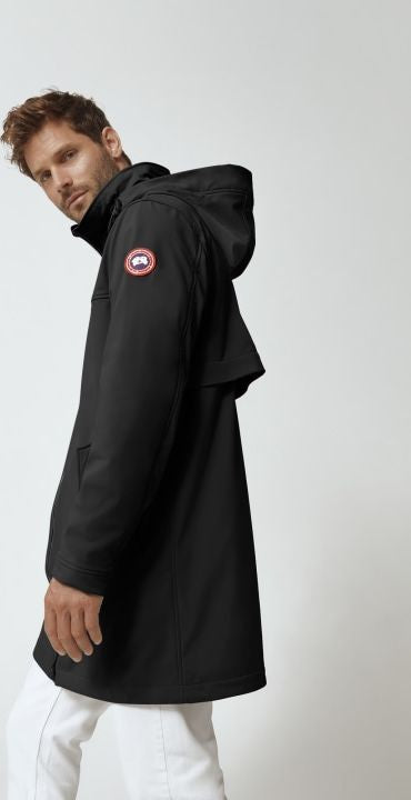 A man wearing a black hooded coat with a round logo on the left arm, standing sideways.