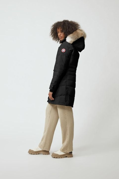 A woman poses in a black parka with a fur-trimmed hood, beige pants, and chunky beige shoes.