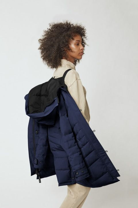 Person with curly hair, wearing a beige sweater and pants, carries a navy blue puffer jacket on their shoulder.