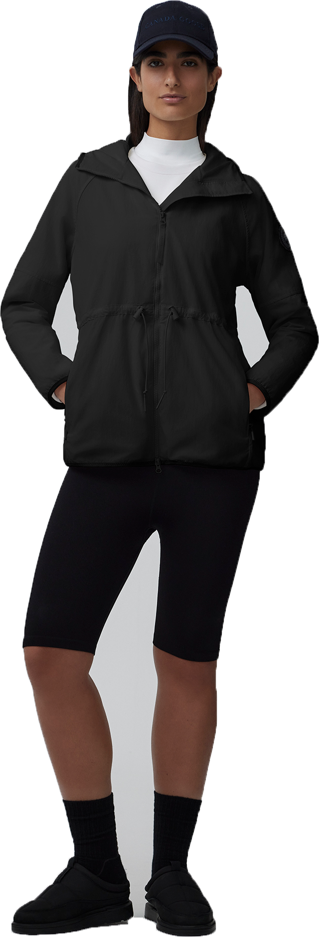 A woman wearing a baseball cap, white turtleneck, a black jacket, black short leggings, and black socks with black shoes.