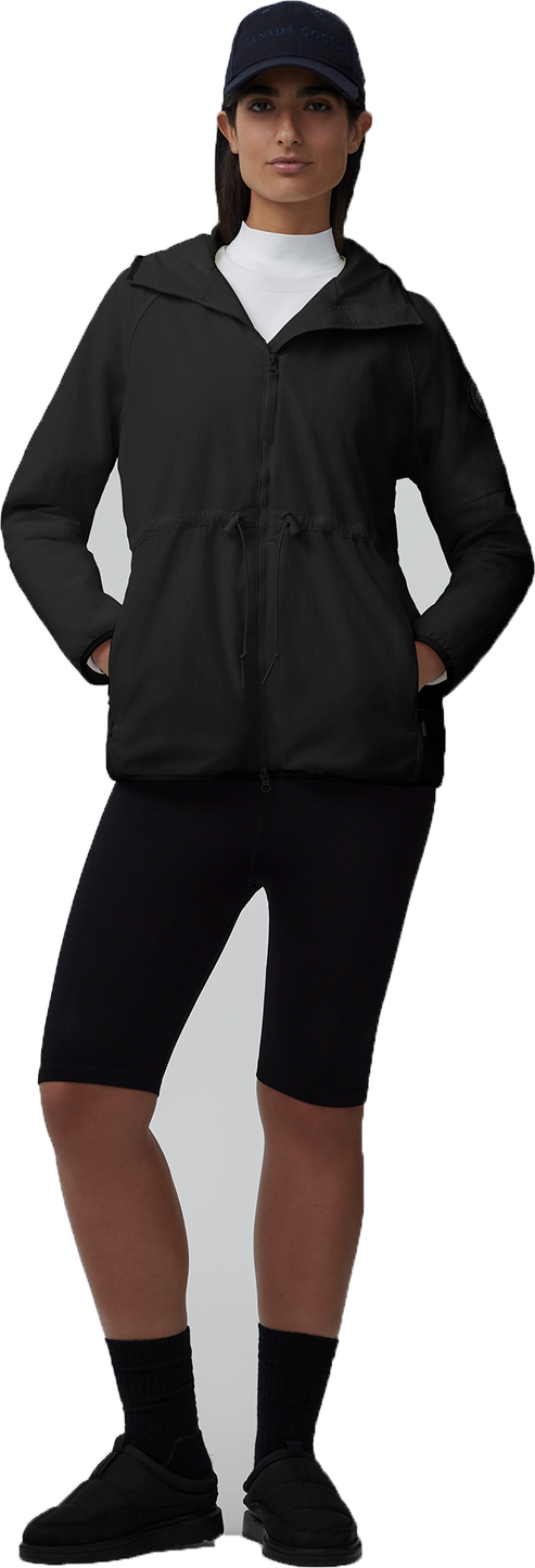 A woman wearing a baseball cap, white turtleneck, a black jacket, black short leggings, and black socks with black shoes.