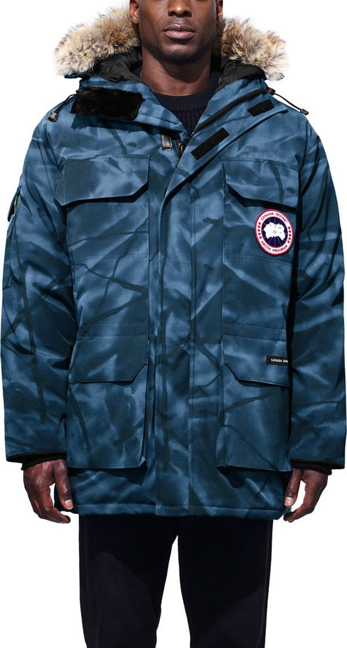Man wearing a blue camo parka with fur-lined hood and the circular logo on the upper right sleeve.
