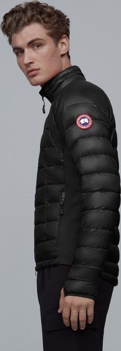 A slender man with curly hair wears a black quilted jacket with a circular red-and-white patch on the upper arm.