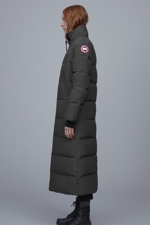 A woman stands wearing a long, quilted dark green coat and black boots.