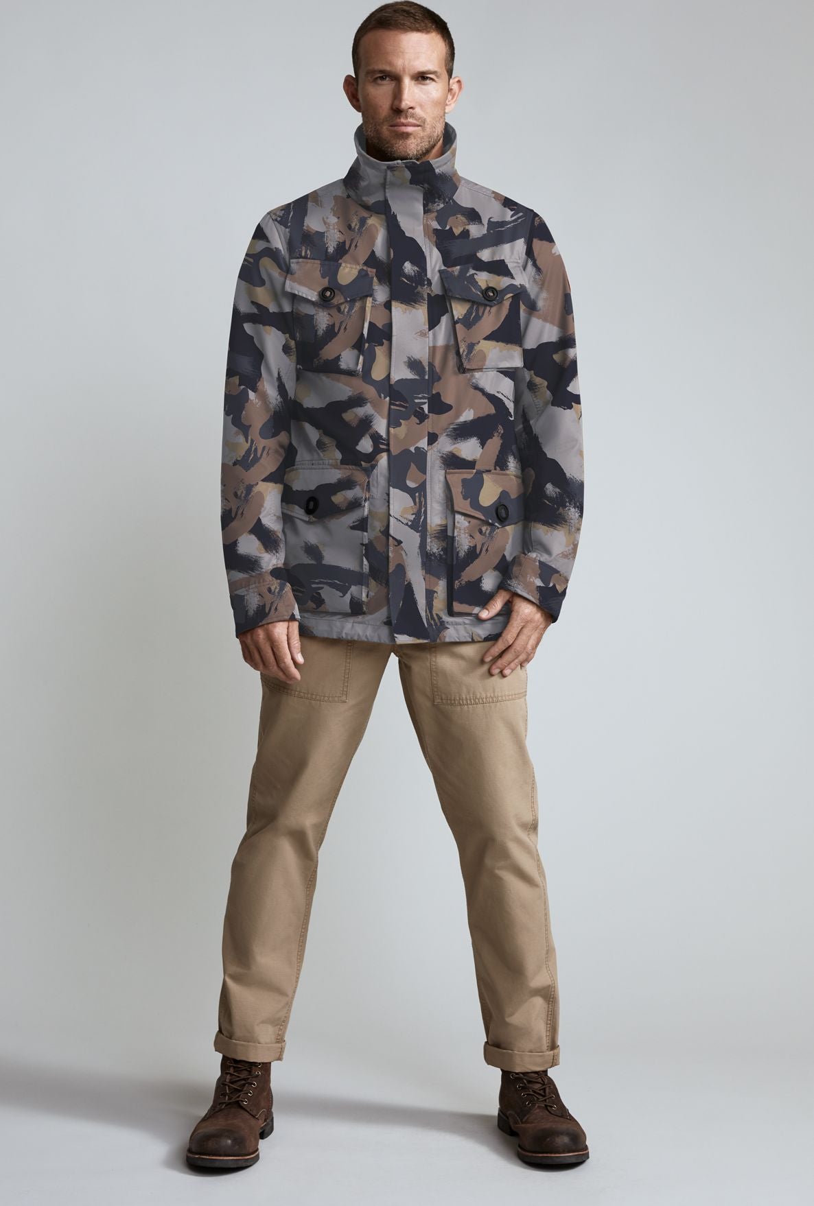 A man in a camouflage jacket, tan pants, and brown shoes stands facing forward.