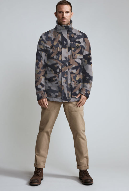 A man in a camouflage jacket, tan pants, and brown shoes stands facing forward.