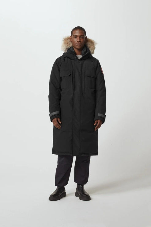 A man wearing a long, black winter coat with a fur-trimmed hood, black pants, and black shoes.