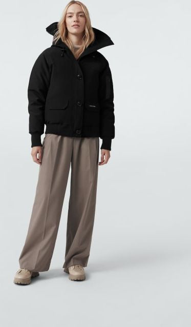 A woman wears a black bomber jacket with two front pockets and beige pants.