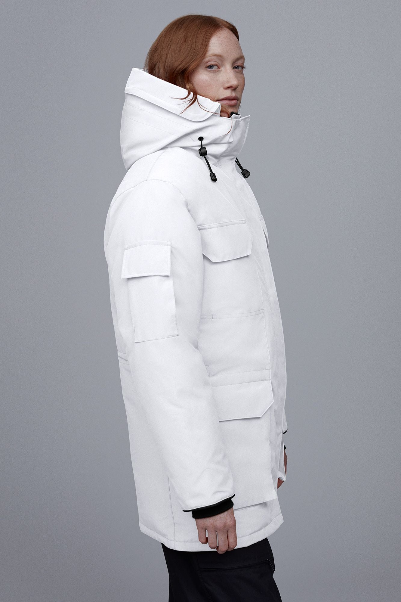 A person is standing in profile wearing a white parka with large pockets and a hood.
