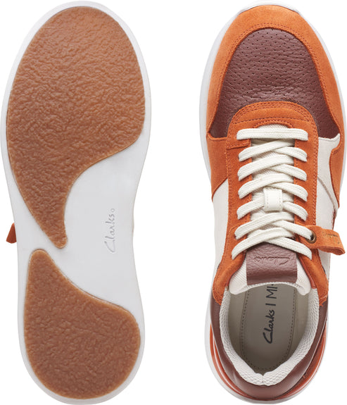An overhead product shot displays the sole and upper portion of a multicolored sneaker. The sole is white and shows a section of the bottom as a light brown. The shoe is colored with off-white panels between brick red and orange suede panels, and white laces.