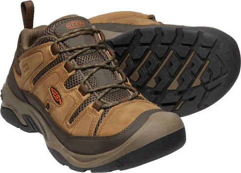 Two light brown hiking shoes, one on its side with a black lug sole, the other standing up, with brown laces and orange accents.