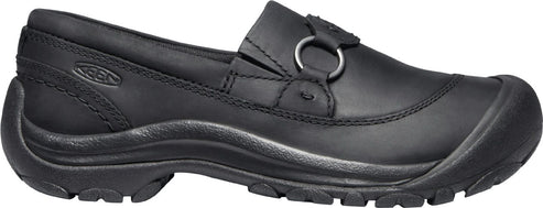 A black slip-on shoe with a rubber sole and a round metal ring on the upper.