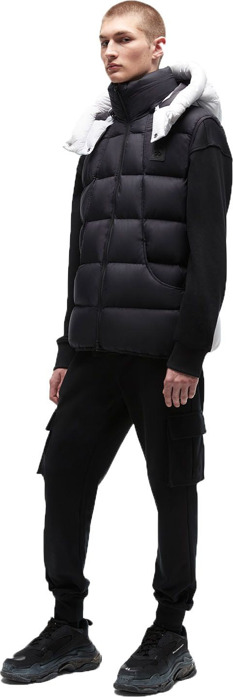 A man in a black puffer vest with a white hood, a black turtleneck, black cargo pants, and black sneakers.