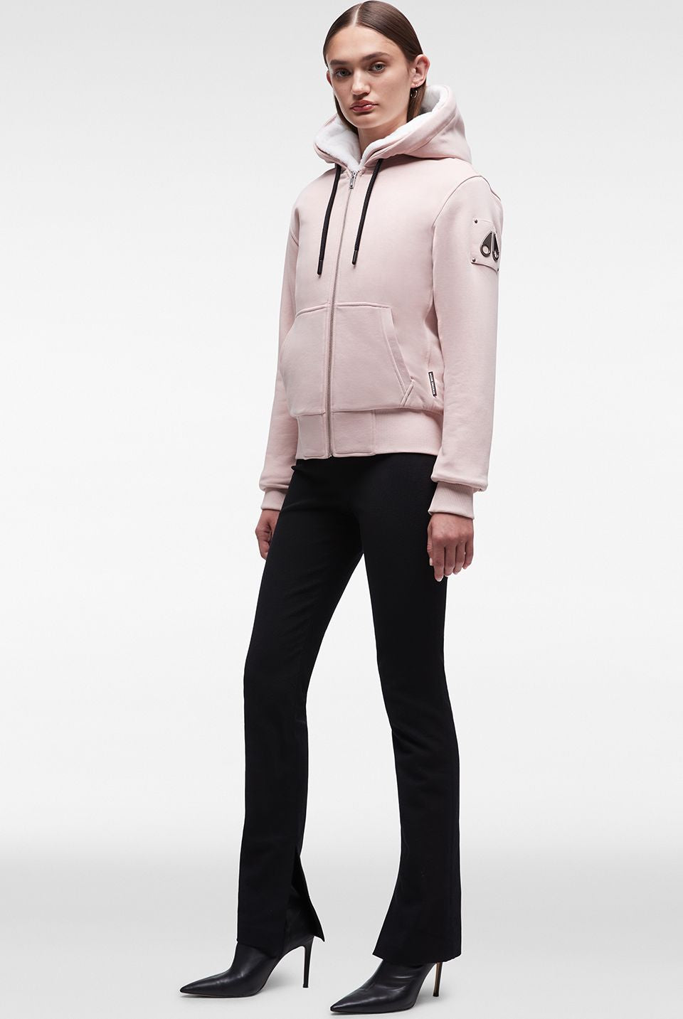 A woman wearing a light pink hoodie and black pants, standing. The hoodie has a light-colored lining inside the hood and a logo patch on the left sleeve. The black pants are fitted and have a slight flare at the bottom, paired with pointy black heels.
