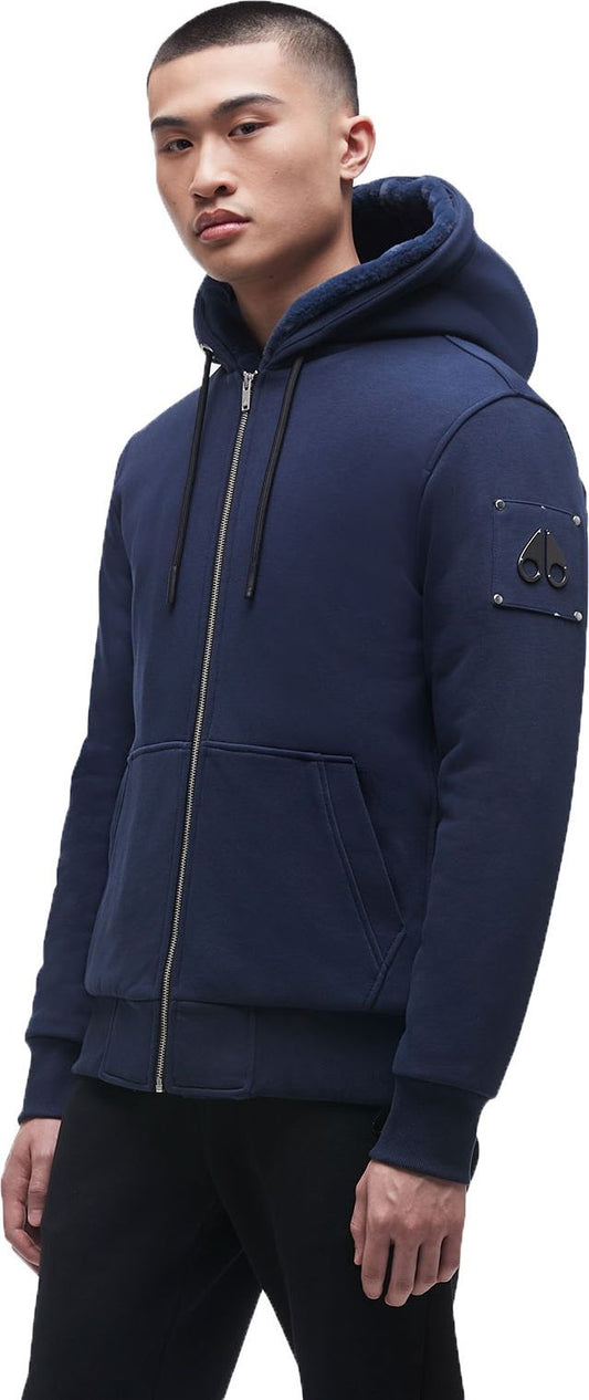 A man wears a blue, hooded zip-up sweatshirt with a logo on the left sleeve and black pants.