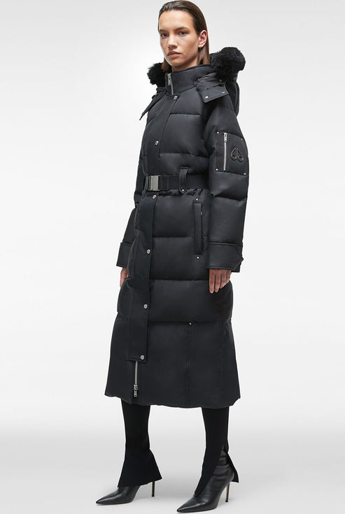 A person wearing a full length black, quilted puffer coat, with a belt at the waist and fur around the hood.