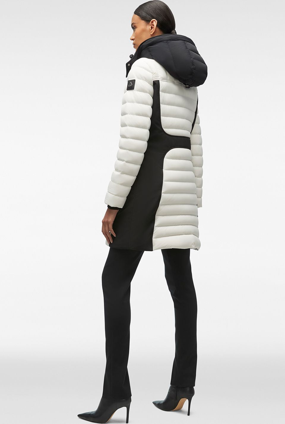 A woman stands in profile wearing a white down coat with black accents and high-heeled black boots.