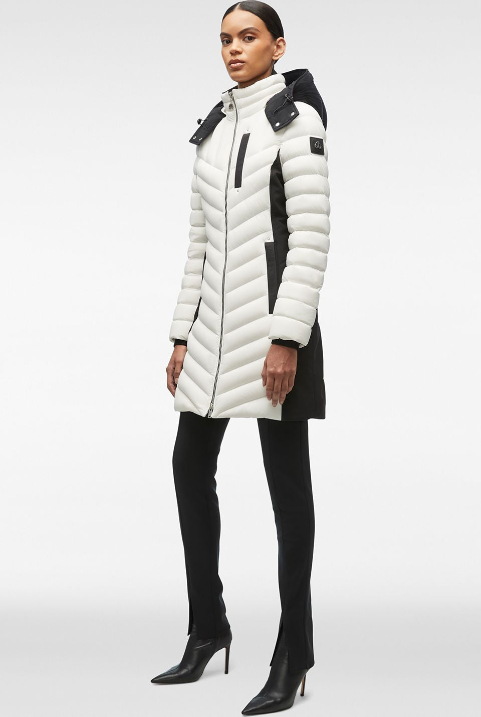 A woman stands in a white and black chevron quilted puffer jacket, black pants, and black pointed-toe stilettos.