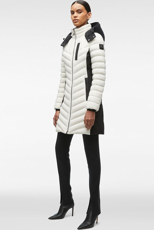 A woman stands in a white and black chevron quilted puffer jacket, black pants, and black pointed-toe stilettos.