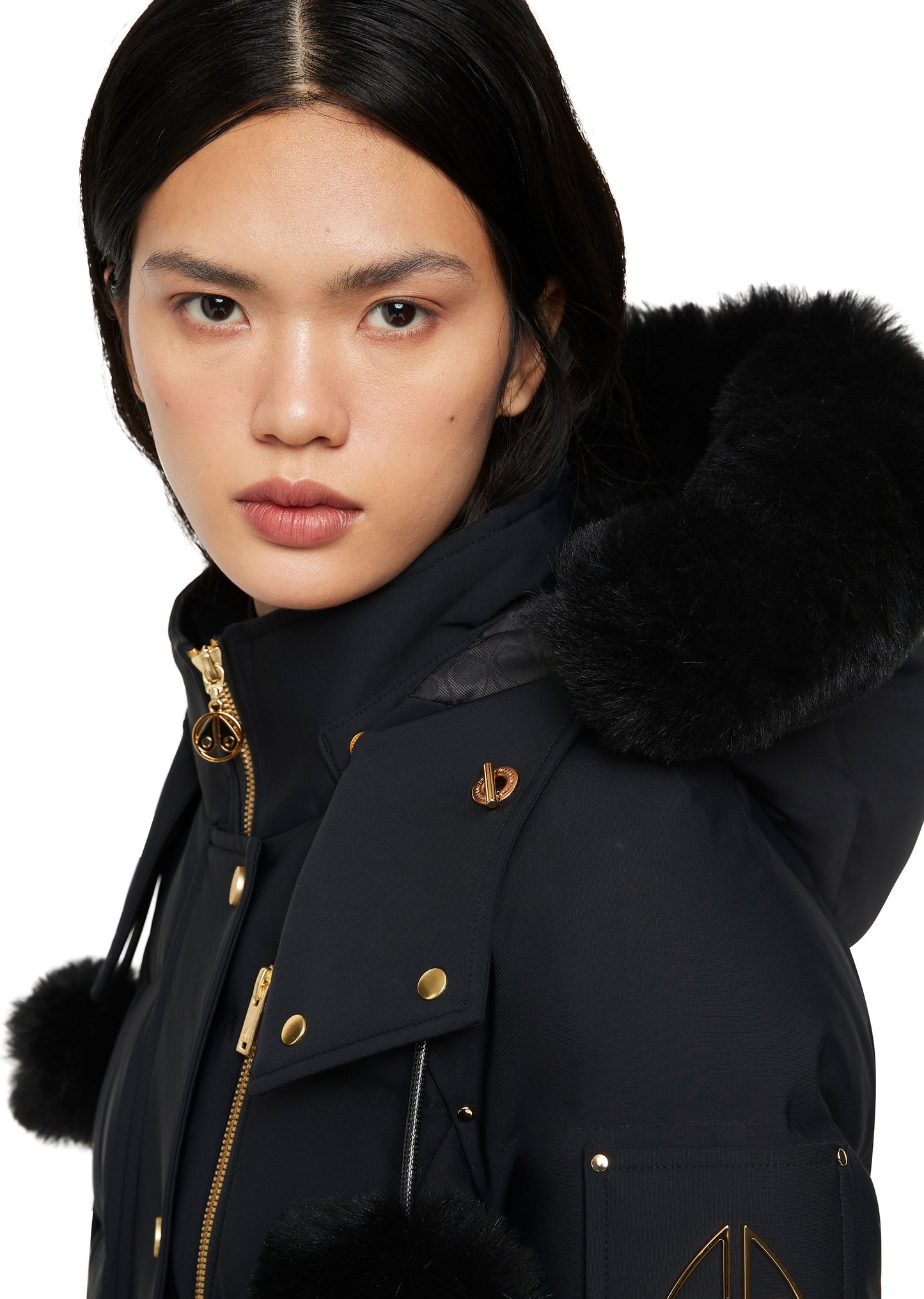 Person wearing a black jacket with fur-trimmed hood and cuffs, gold hardware, and pom-poms.