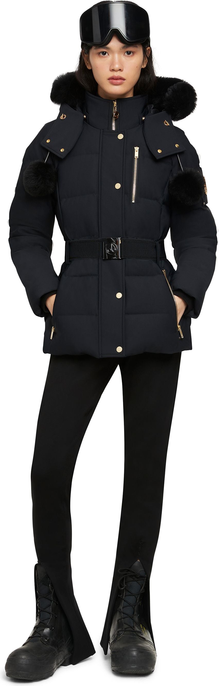 A woman wearing a black ski jacket, black leggings, and black snow boots, also wearing ski goggles on her head.