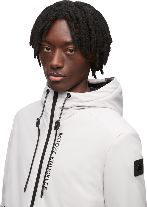 Person wearing a grey hooded anorak with a zipper and a black logo on the sleeve and the words MOOSE KNUCKLES on the chest.