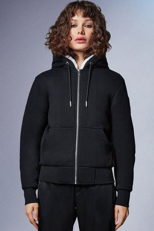 A woman with curly brown hair wearing a black zip-up hoodie with a white sherpa-lined hood and black pants.