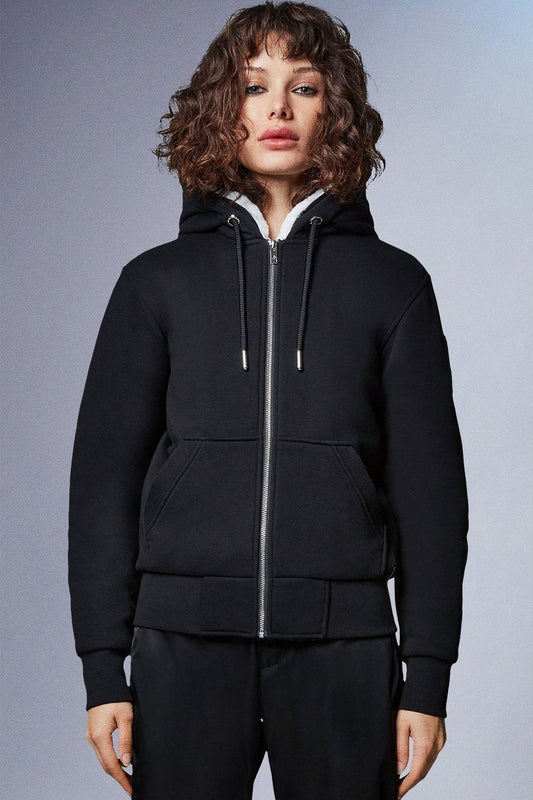 A woman with curly brown hair wearing a black zip-up hoodie with a white sherpa-lined hood and black pants.