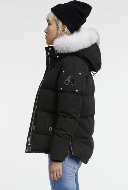 A woman wearing a black puffer coat with a white fur-trimmed hood, a black beanie, and light blue jeans.