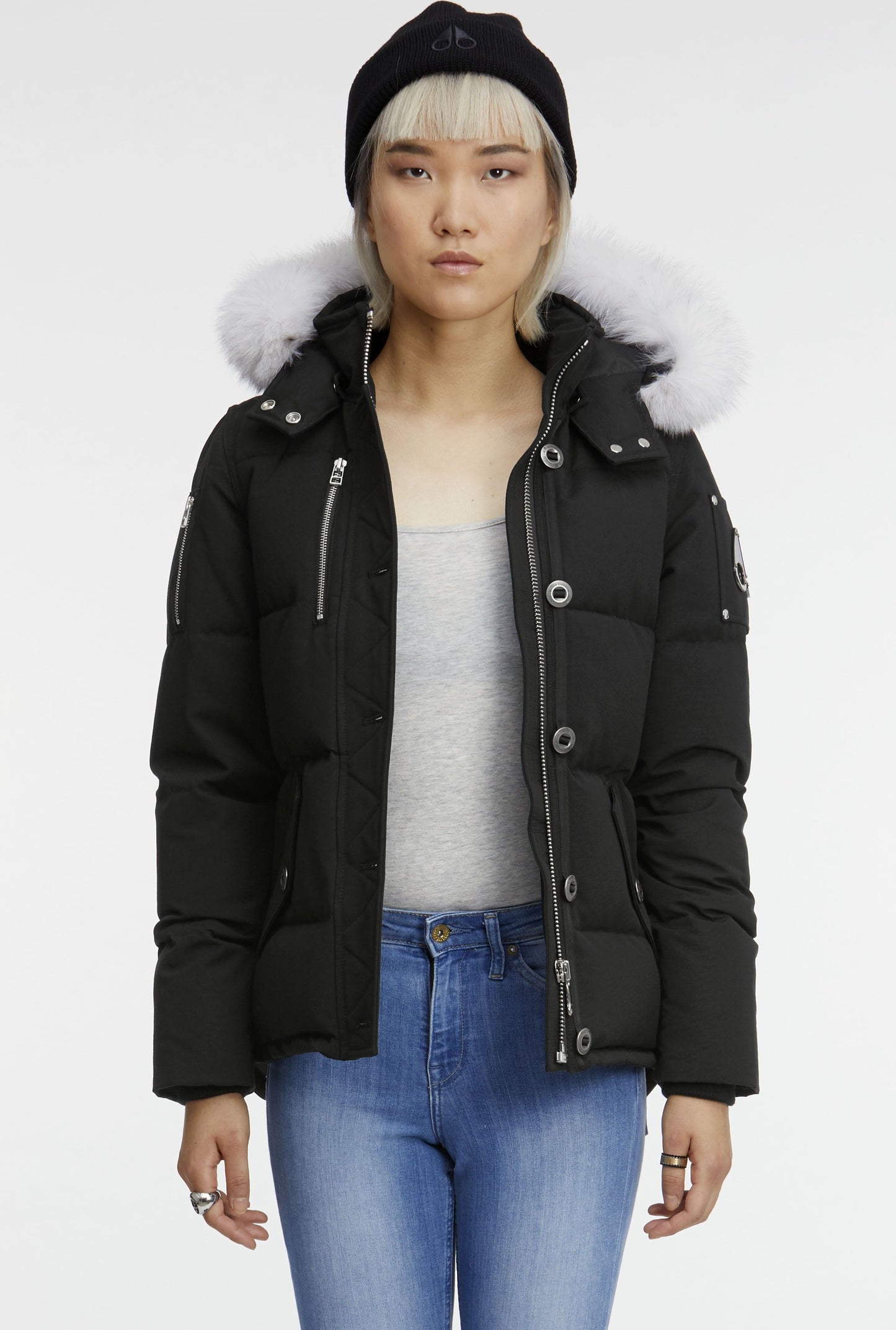 A woman wearing a short, black puffer coat with a fur-trimmed hood, a black beanie, a gray shirt, and blue jeans.