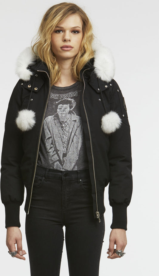A model is wearing a black bomber jacket with white fur around the hood and white fur pom-poms.