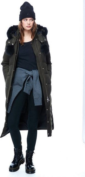 A person wearing a black beanie, a long olive-green parka with fur trim on the hood, a black shirt, black leggings, and black platform boots.