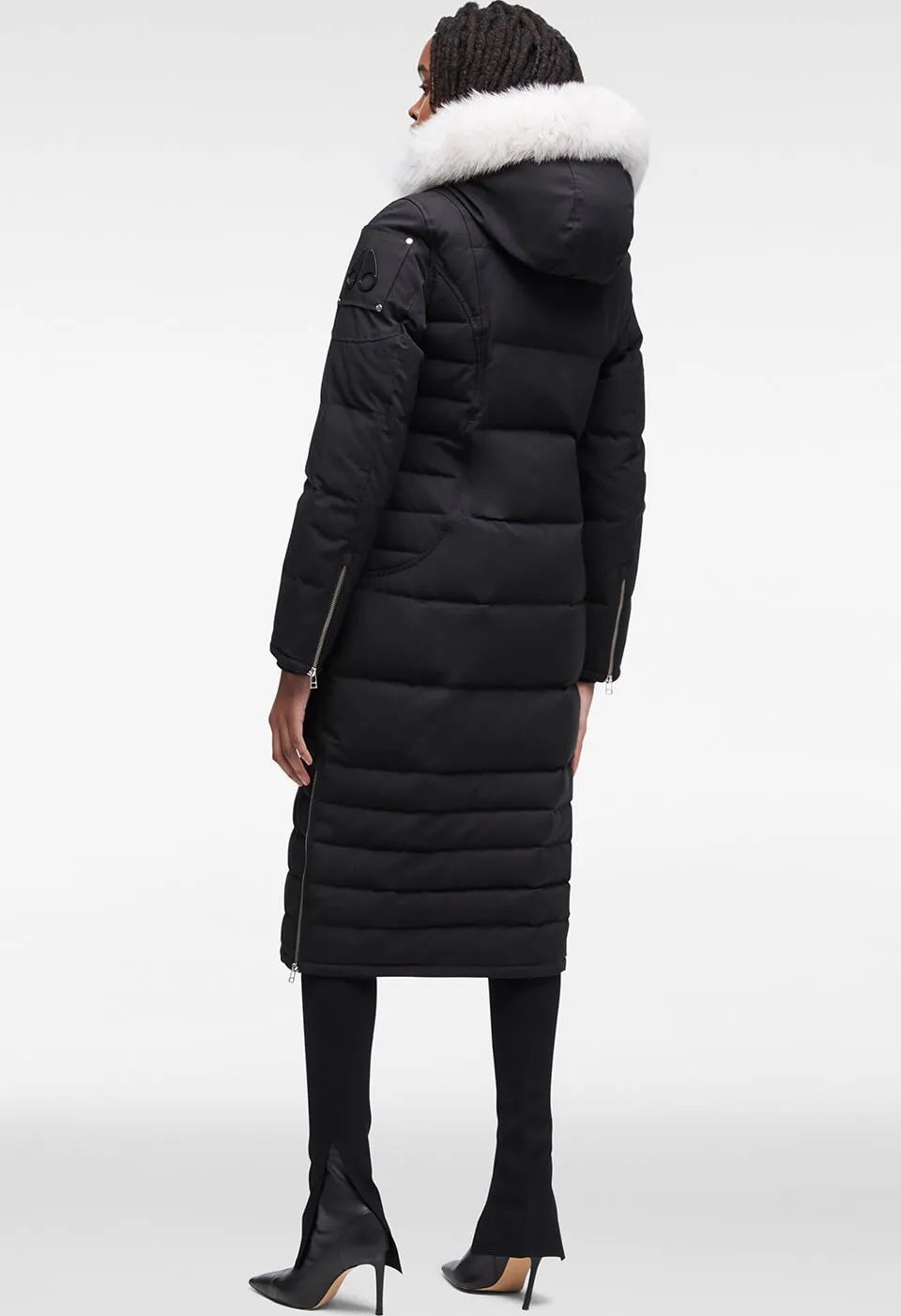 A woman standing and wearing a long black puffer coat with white fur trim around the hood, black leggings, and black stiletto boots.