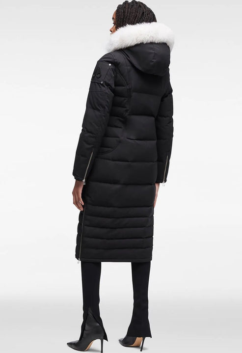 A woman standing and wearing a long black puffer coat with white fur trim around the hood, black leggings, and black stiletto boots.