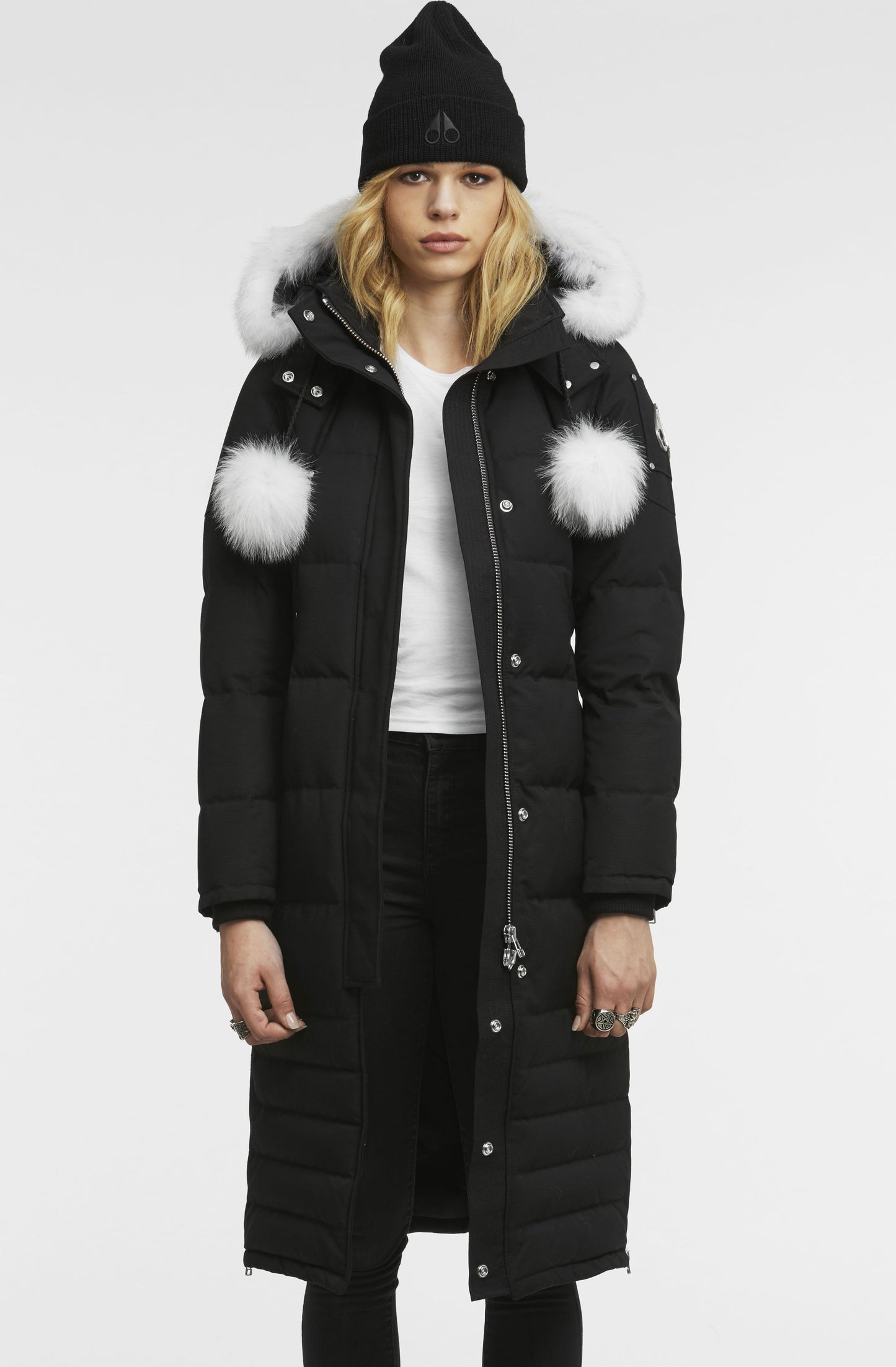 A woman wears a long black down coat with a white fur-lined hood and white puff balls on the shoulder.