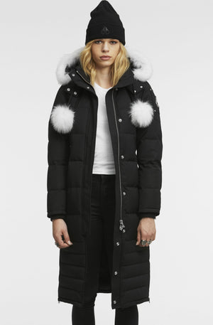 A woman wears a long black down coat with a white fur-lined hood and white puff balls on the shoulder.
