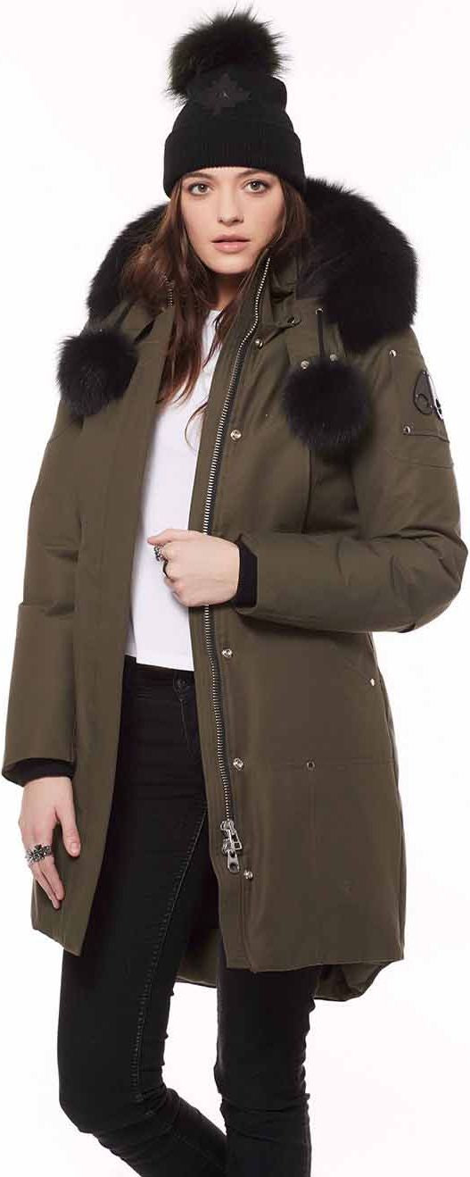 A woman wearing an olive green coat with black fur trim, a white shirt, and a black beanie.