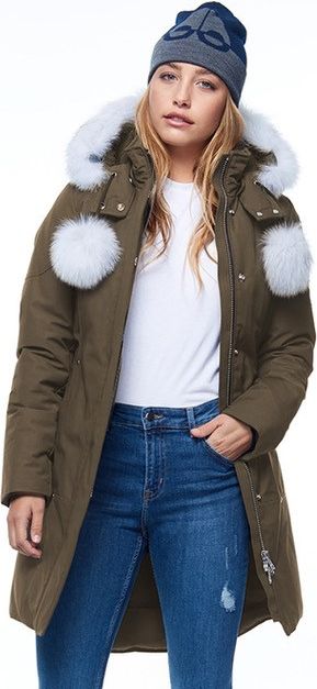 A woman wearing a beanie, an olive green coat with white fur trim, a white shirt, and blue jeans.