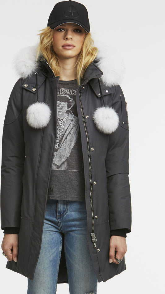 A person wearing a long, dark coat with white fur trim and pom poms, paired with a gray t-shirt featuring a black-and-white graphic, blue jeans, and a black baseball cap.