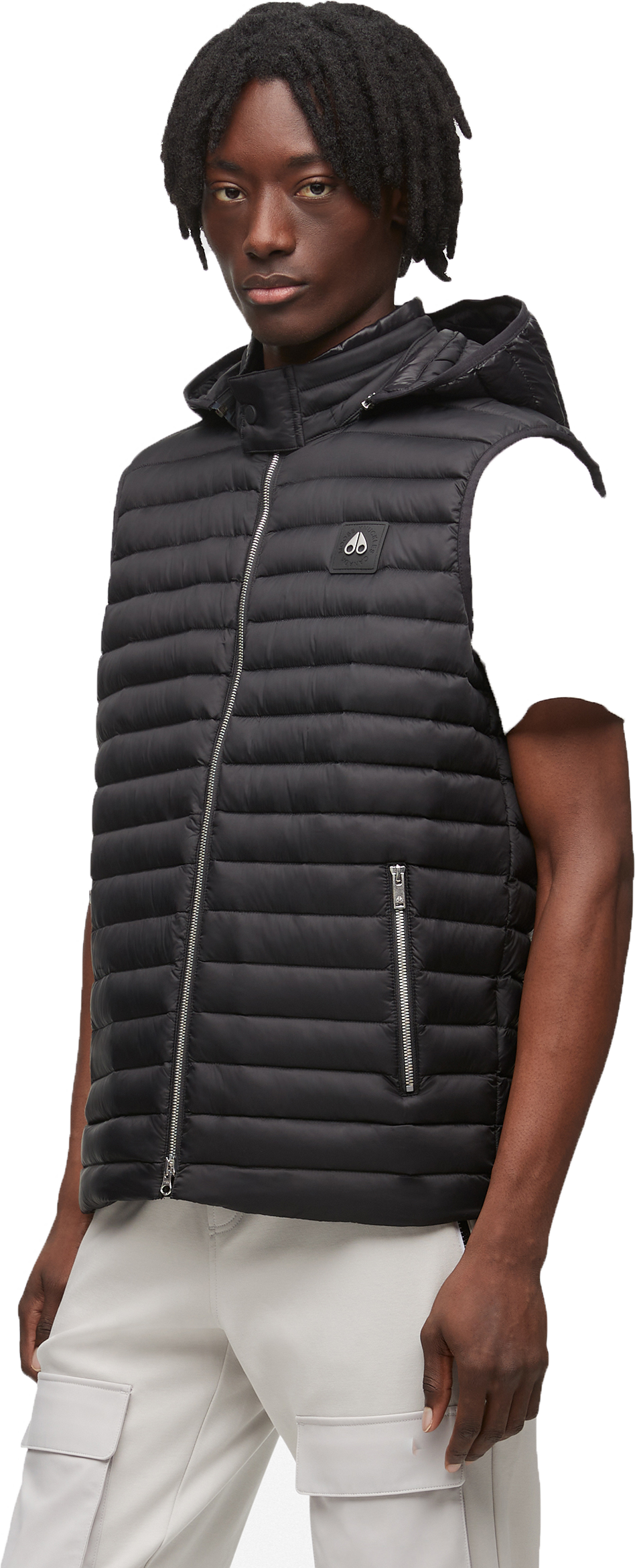 A Black person with dark hair wears a black hooded vest and light-colored pants.