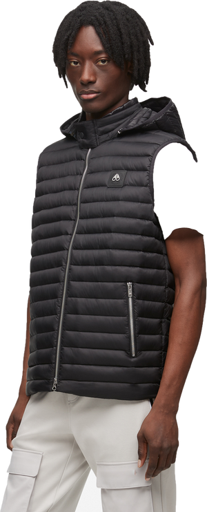 A Black person with dark hair wears a black hooded vest and light-colored pants.