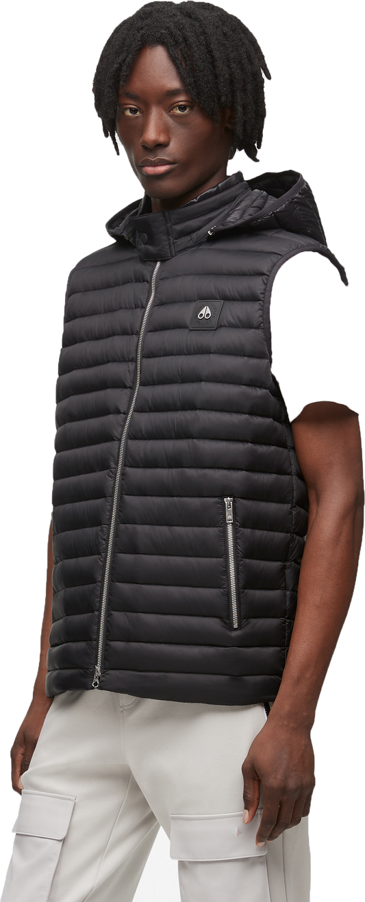 A Black person with dark hair wears a black hooded vest and light-colored pants.
