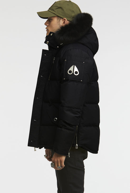 A man wearing a black puffer coat with a fur-trimmed hood and metal logo detail on the arm.