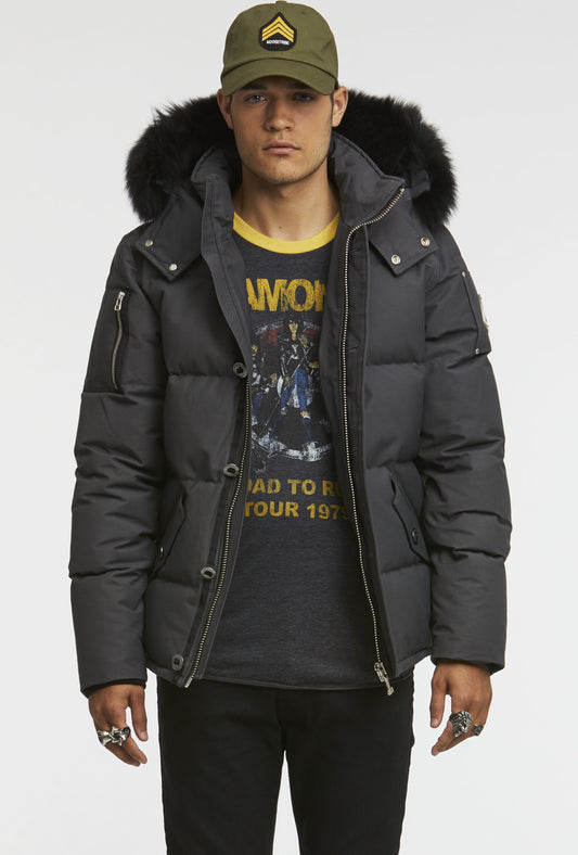 A man wears a gray puffer jacket with a fur-trimmed hood, a Ramones t-shirt, black pants and a green cap.
