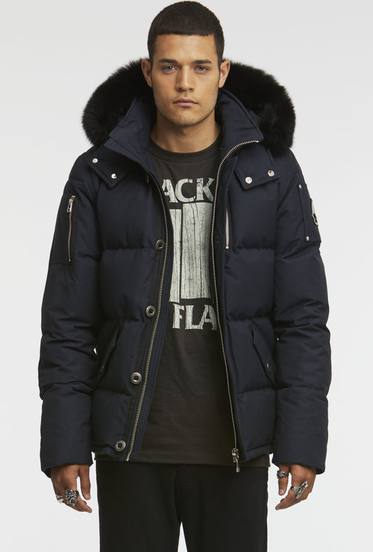 A man wearing a navy blue puffer jacket with a black fur-trimmed hood over a black t-shirt with a graphic.